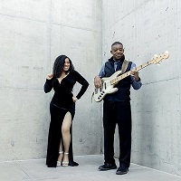 a woman and her father standing against a blank wall, the father is playing the guitar