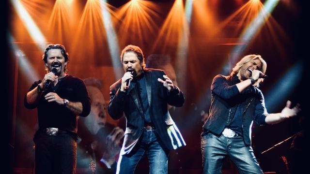 three white men singing on stage with jeans on and dark shirts with orange spotlights behind them