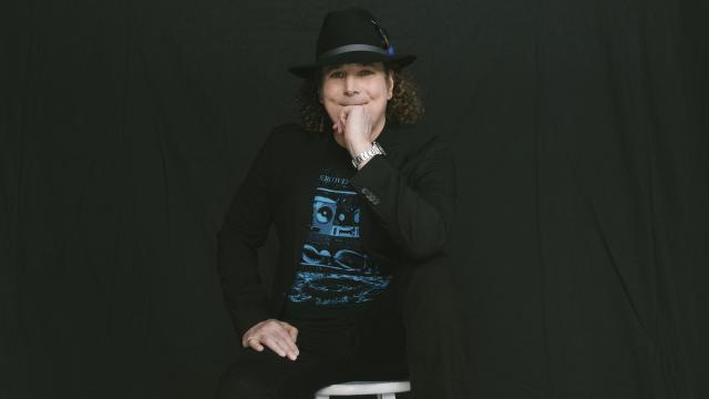 white man with curly brown hair wearing all black and a black hat in front of a black background.