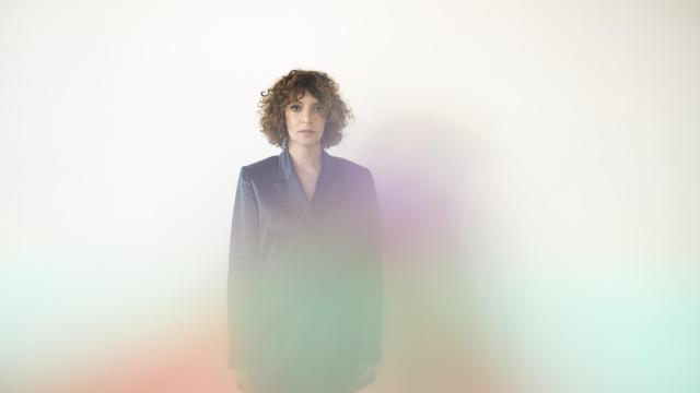 Woman with lighter skin and brown curly hair standing in the center, looking at the viewer. There is a rainbow like fog in front
