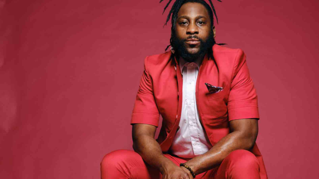 Michael Mwenso, a man with long dreadlocks in a ponytail, sits in front of a red wall in a red suit