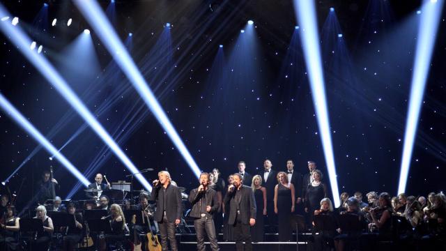 Three men with a chorus behind them standing on a dark stage singing