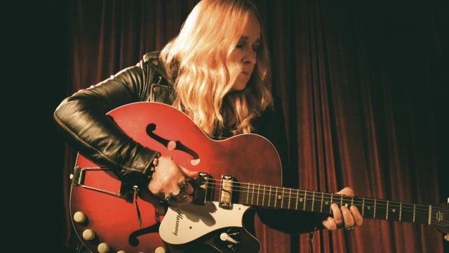 Blonde woman holding a red guitar looking to the right with a dark background