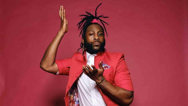 A man with dreadlocks in a ponytail wears a red suit, sitting on a chair in front of a red background. He looks at the camera.