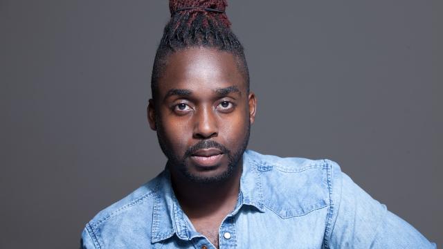 A man with a bun on top of his head looks at the camera, wearing a denim shirt