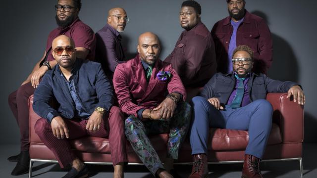 7 black men sitting around a maroon couch looking at the viewer