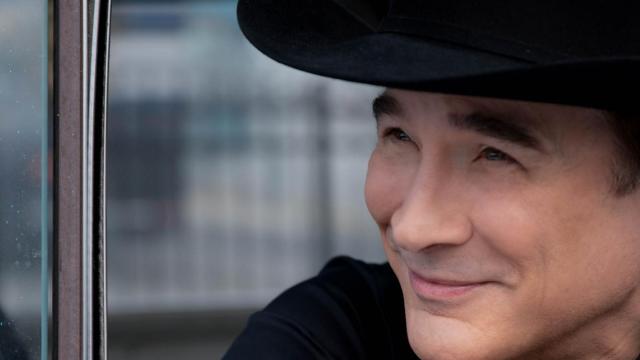 Clint Black smiles off camera as he leans out a car door. He wears a black shirt and black cowboy hat. 