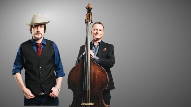 two men standing next to each other, both white, one wearing a hat and the other holding a bass