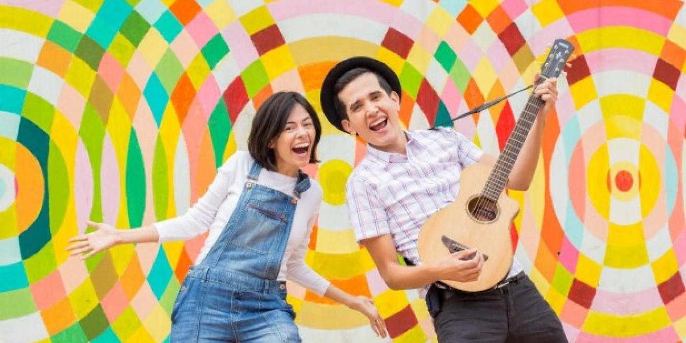 A woman dressed in jean overalls and a man wearing a hat, playing guitar, smile at the camera