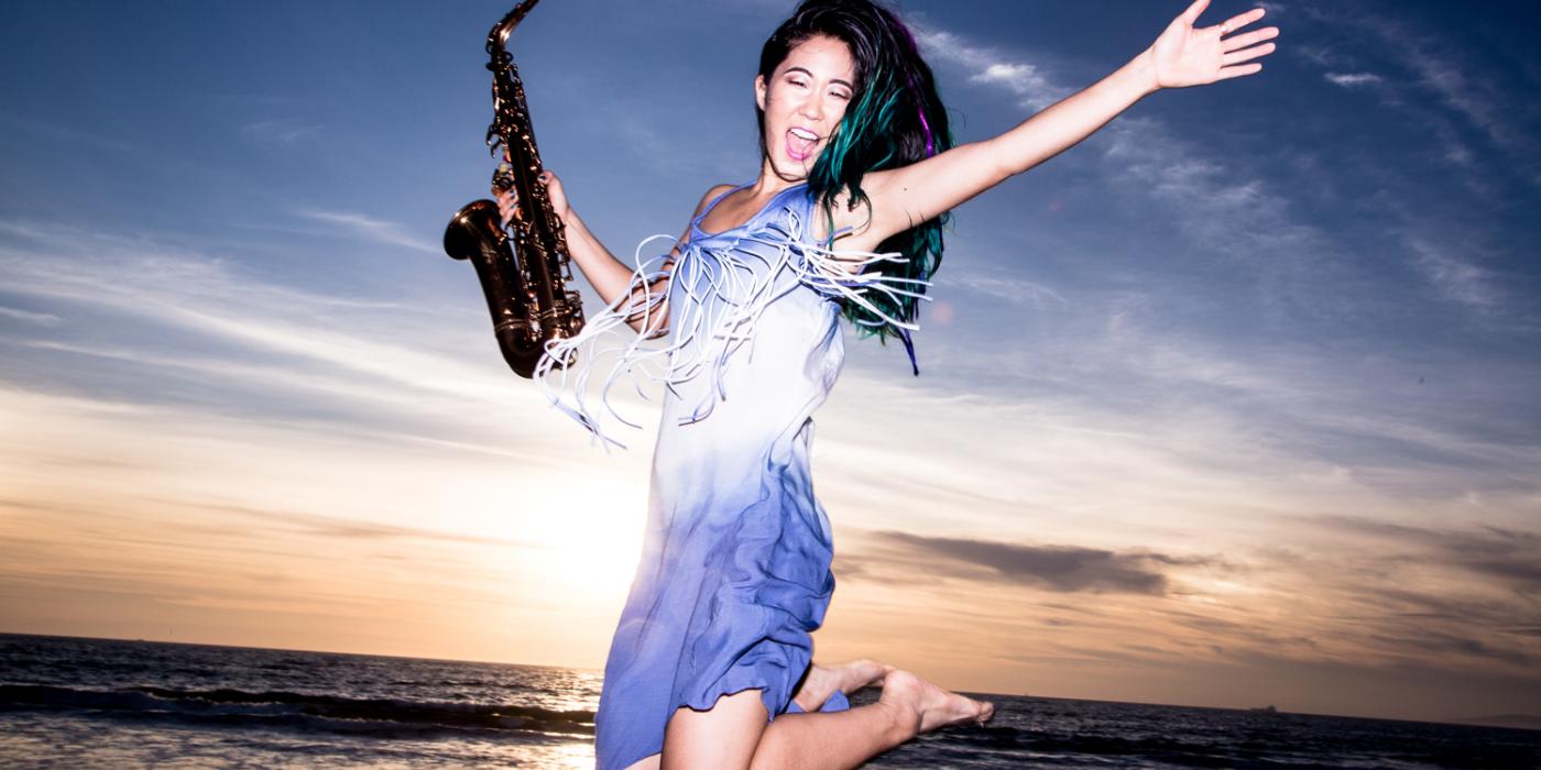 A woman with long black hair is jumping in the air, saxophone in hand, at the beach with a sunset behind her 