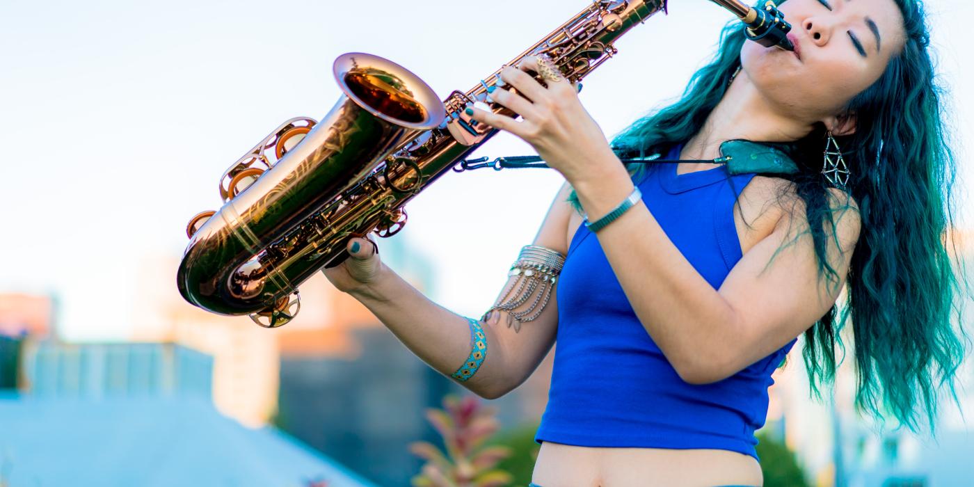 A woman with long wavy green hair, wearing a royal blue shirt, plays a saxophone with eyes closed and her head leaning back