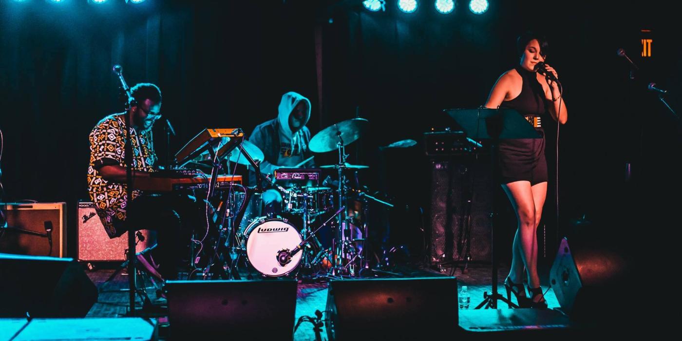 A band plays on a stage with a woman at the microphone, a man playing drums and a man playing guitar