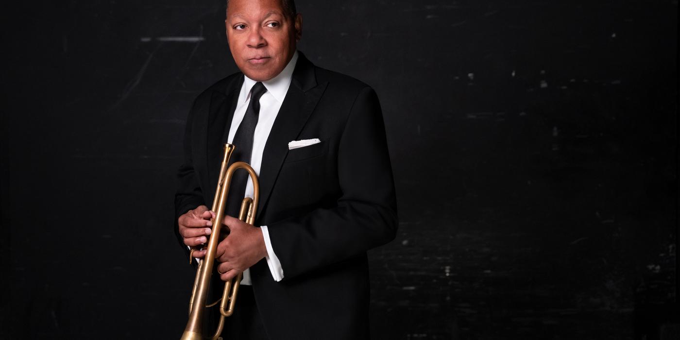 a man in a black suit stands against a black wall with a trumpet