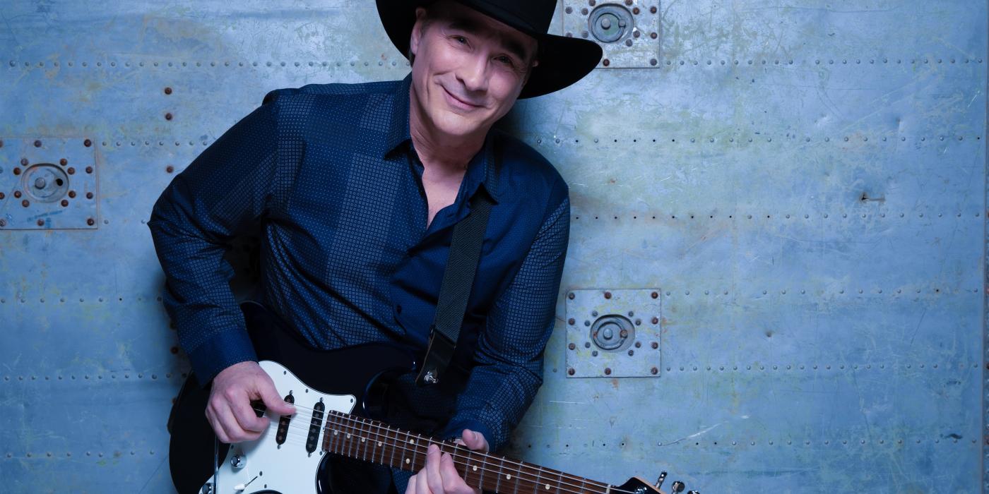 A man in a dark blue shirt stands against a blue wall, electric guitar in hand and a black hat on his head