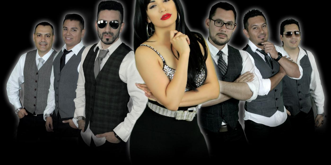 A woman with long black hair and bright red lips, wears all black with a silver studded top. Her hand is under her chin and her hip juts out to one side. Six men, three on either side of her, wear white button up shirts, black ties and vests