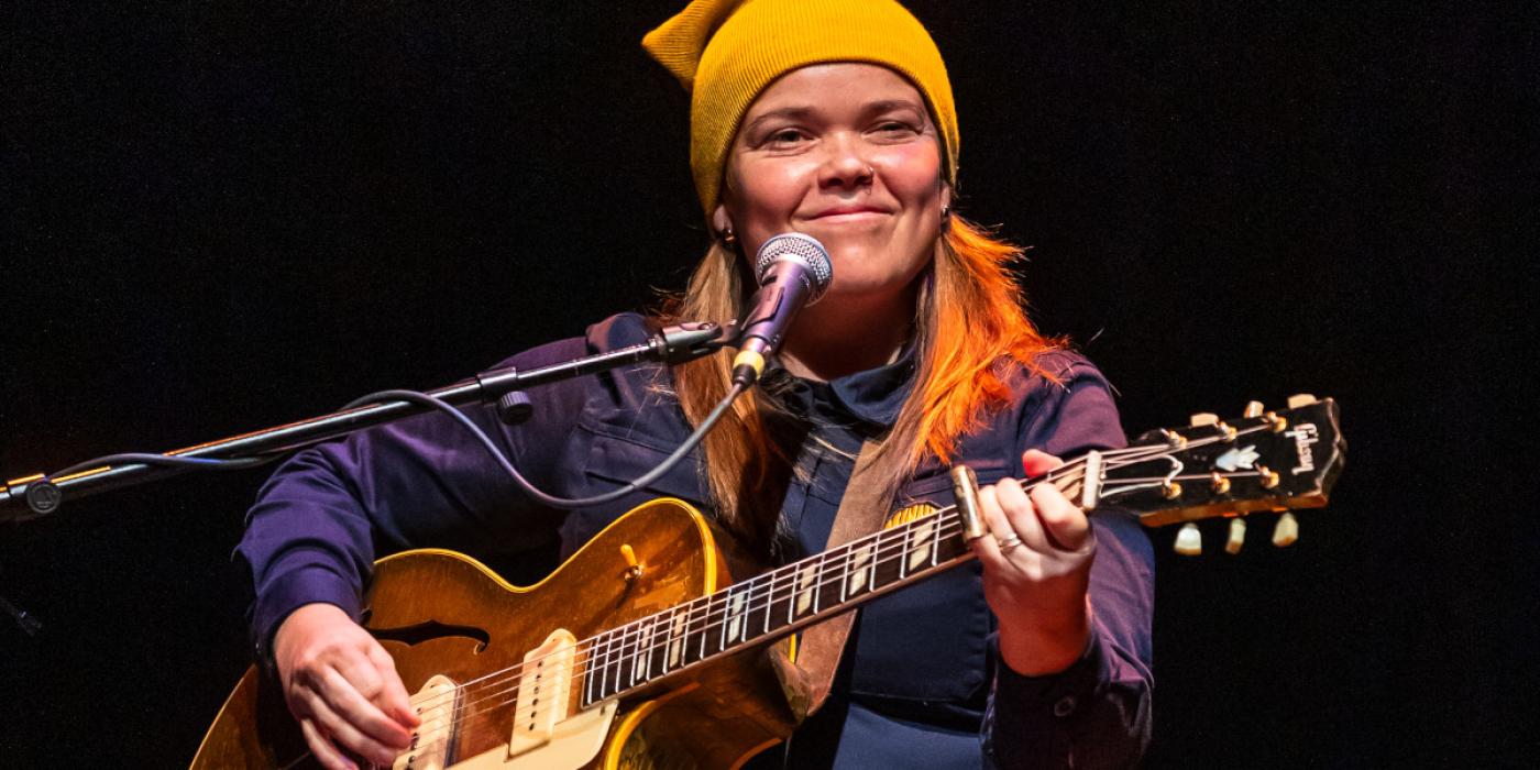 Young tan woman wearing a yellow beanie, playing a guitar on stage.