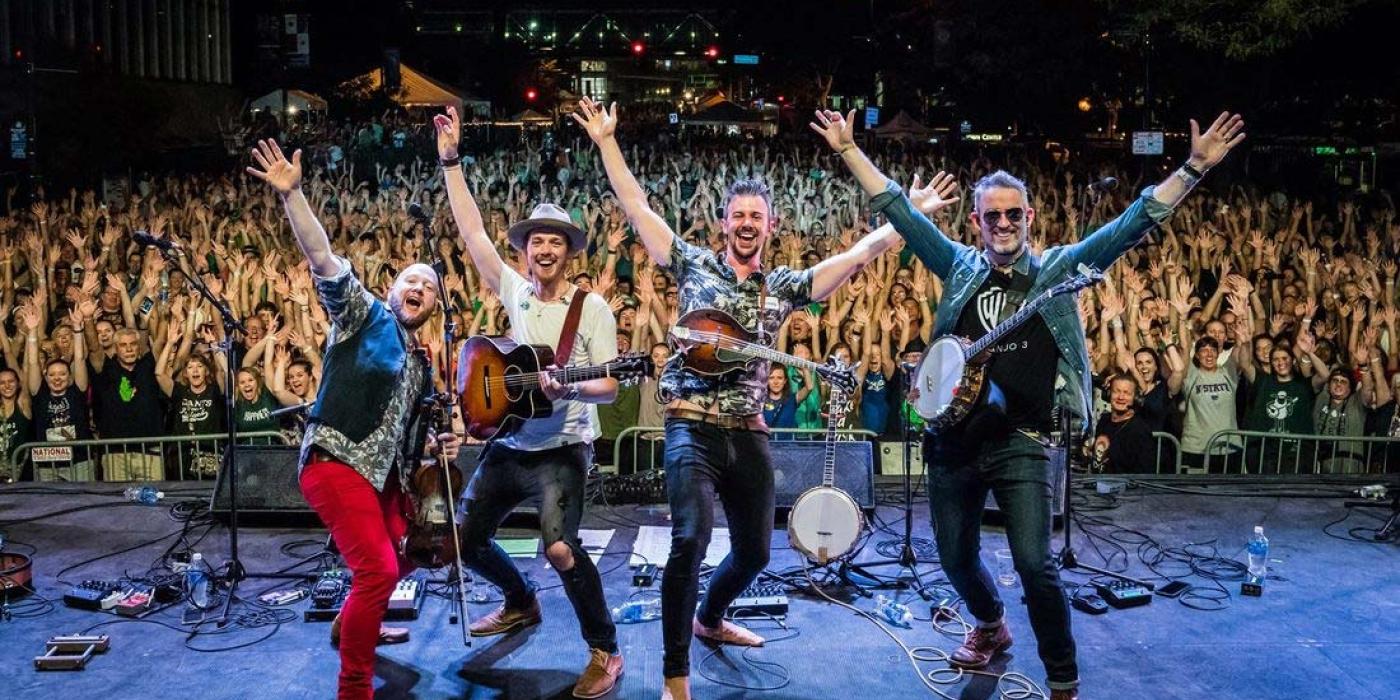  With hands in the air, We Banjo 3 faces the back of the stage with audience in background