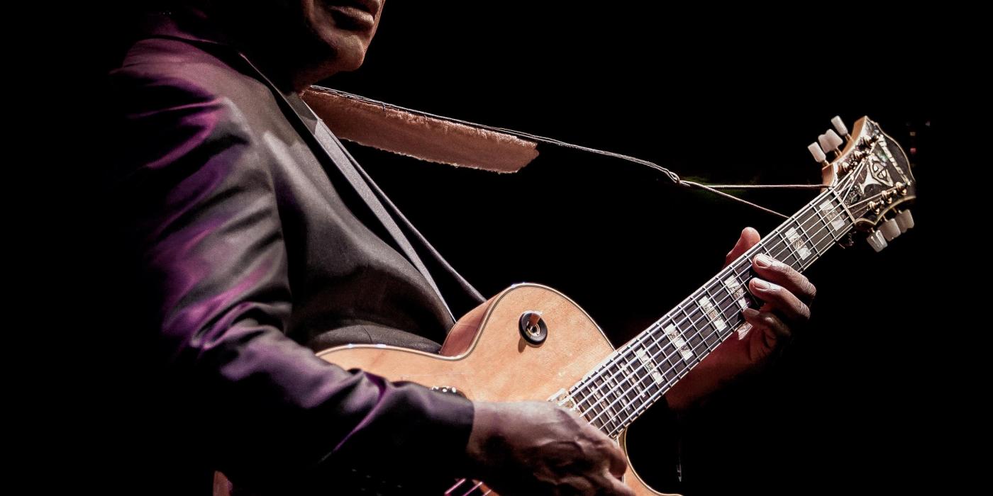 A man with short black hair wears a grey sport coat. He holds an electric guitar, fingers on the fret and strings.