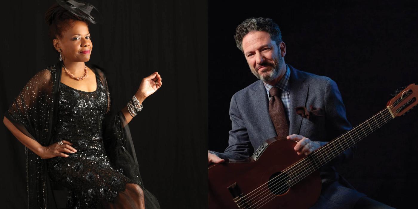 A woman in a black dress sits on a chair, posing with a smile, and a man in a gray suit sits with his guitar