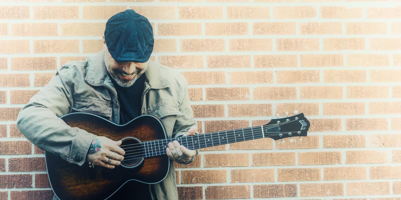 Blaine Long, sporting a beard and wearing a driver cap and khaki jacket leans against a red brick wall with his amber acoustic guitar in his hands