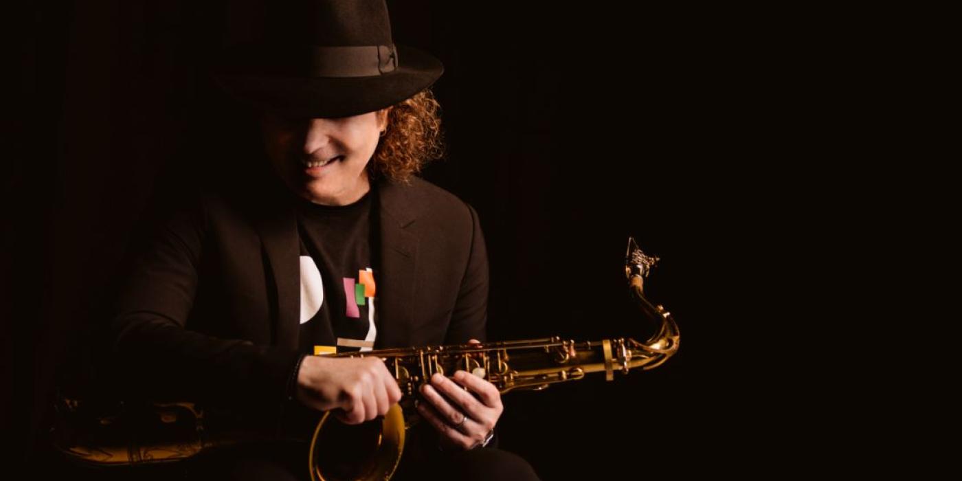 A man with shoulder length curly hair wears all black, blending in with the black background so that you only see his hands holding a saxophone and his face smiling under his fedora.