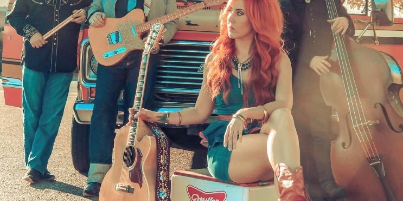 Three men and a woman wear country western wear, holding guitars in front of a pickup truck