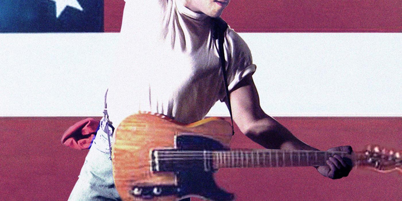 A man wears a white t-shirt with the sleeves rolled up and jeans. He is playing the guitar and leaping with an American flag behind him