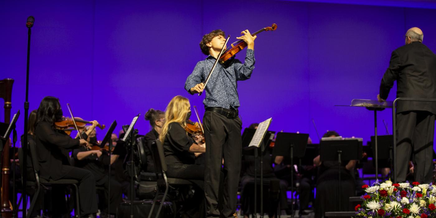 A young man plays the violin on stage with an orchestra behind him