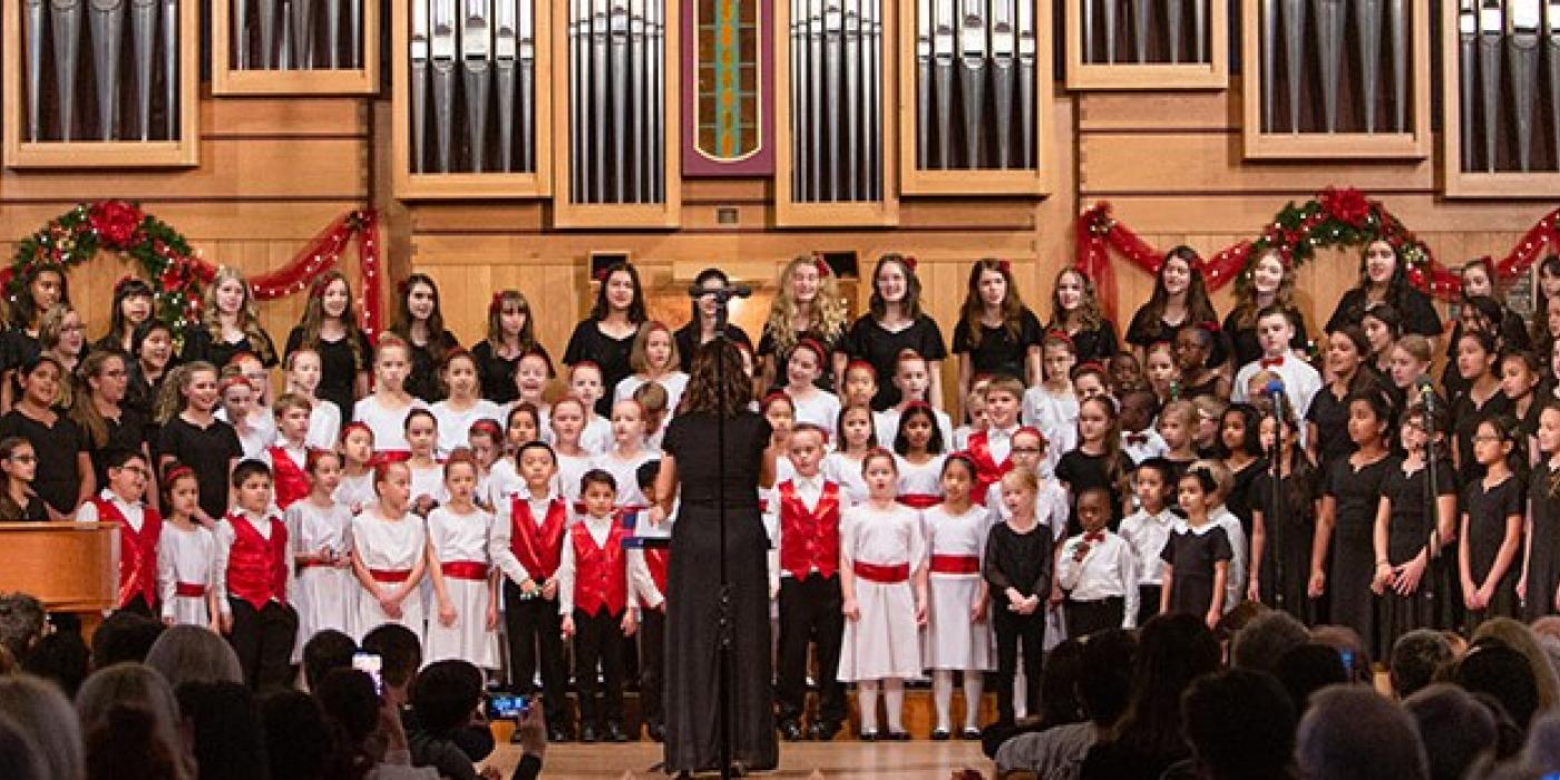 A large group of children stand on risers in front of a church organ as they sing.