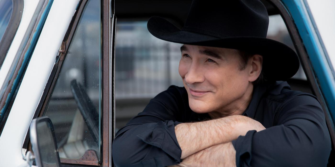 A man leans out the window of a baby blue truck, arms folded and his chin resting on his forearm. He wears a black hat and shirt. 