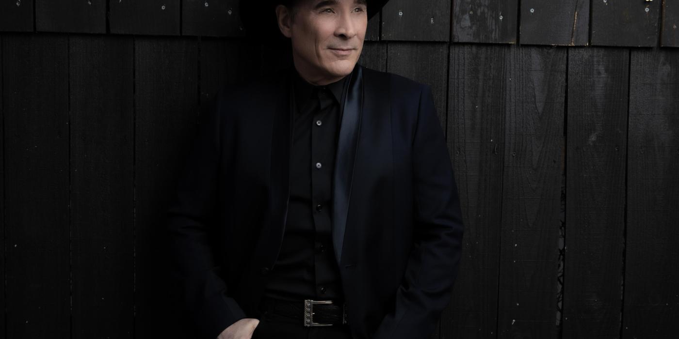 A man all dressed in black - hat, shirt and jeans - leans against a dark brown wall covered in wood slats