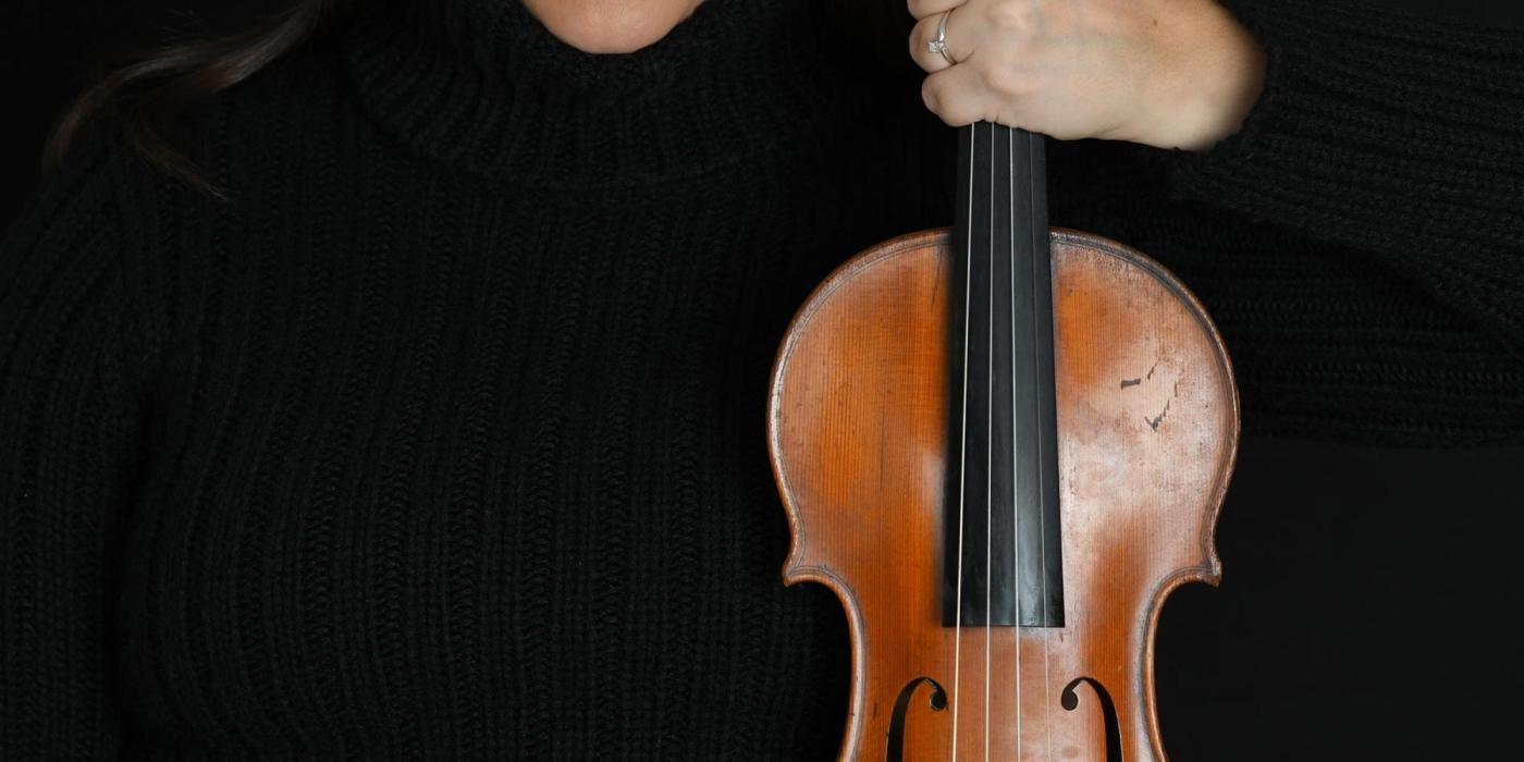 A woman with long dark hair pulled back from her face wears glasses and a black turtleneck sweater. She holds a violin in her left hand. 