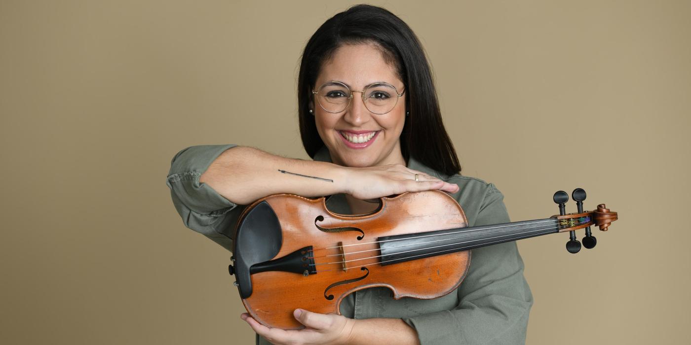 A woman with long black hair and glasses faces the camera holding her violin between her arms