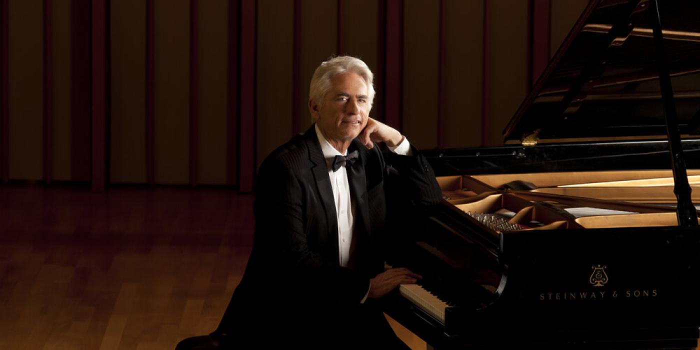 David Benoit - a man with silver hair - sits at a grand piano in a tuxedo looking at the viewer with a smile