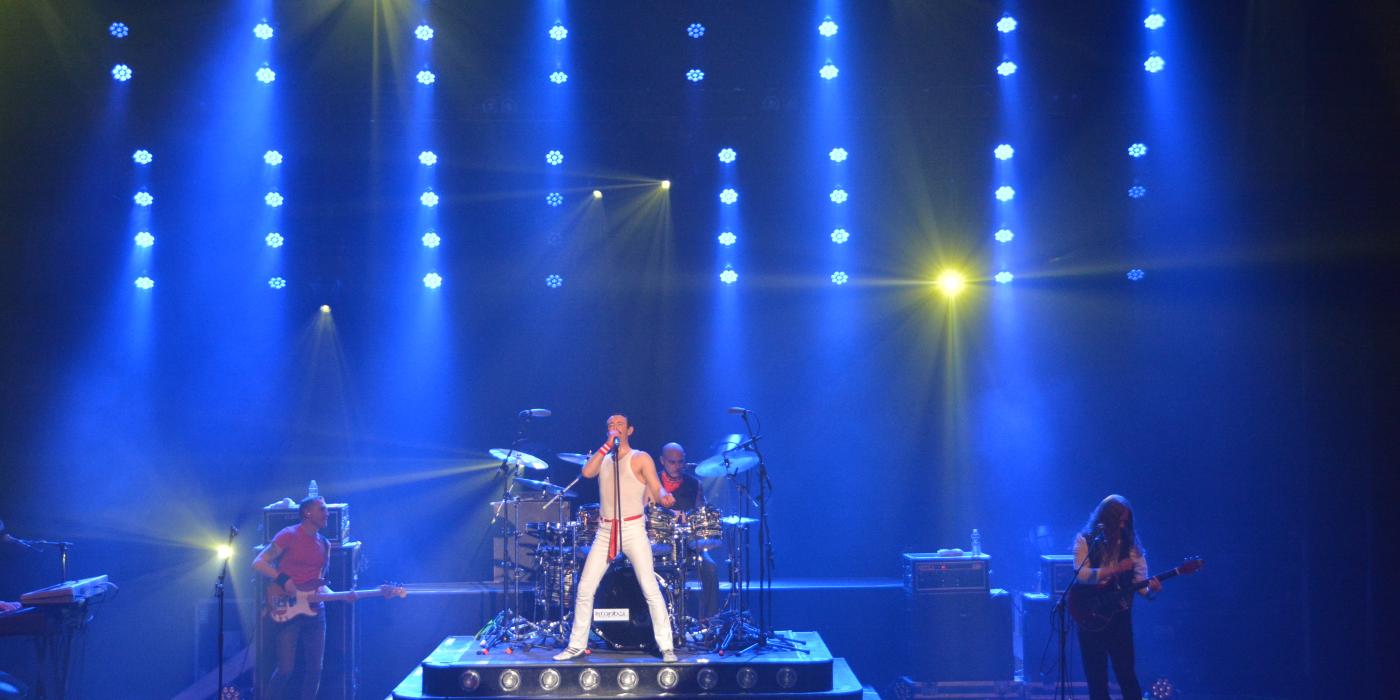 A man dressed in white jeans and a white sleeveless shirt stands center stage with blue lights streaming behind him