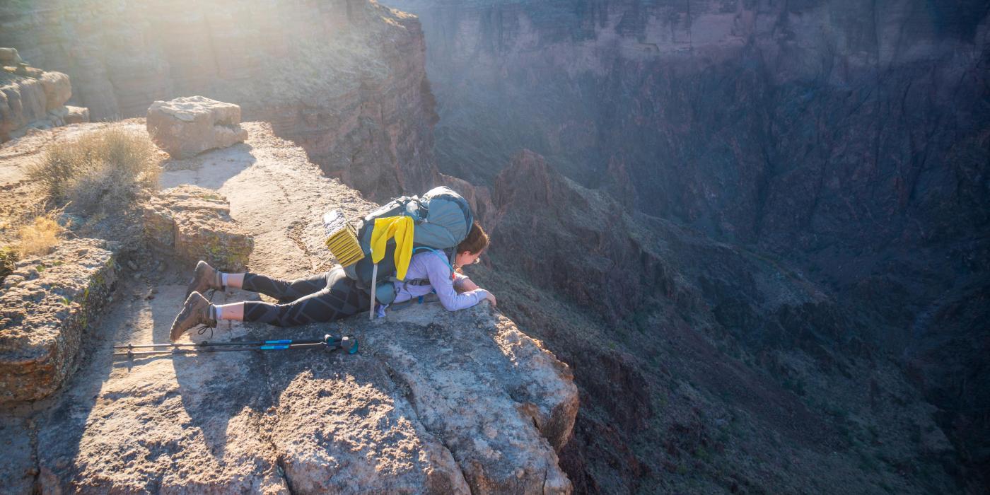 A person lays on their stomach looking over the edge of a rocky cliff, down into a canyon where the sun appears to be setting in the distance.