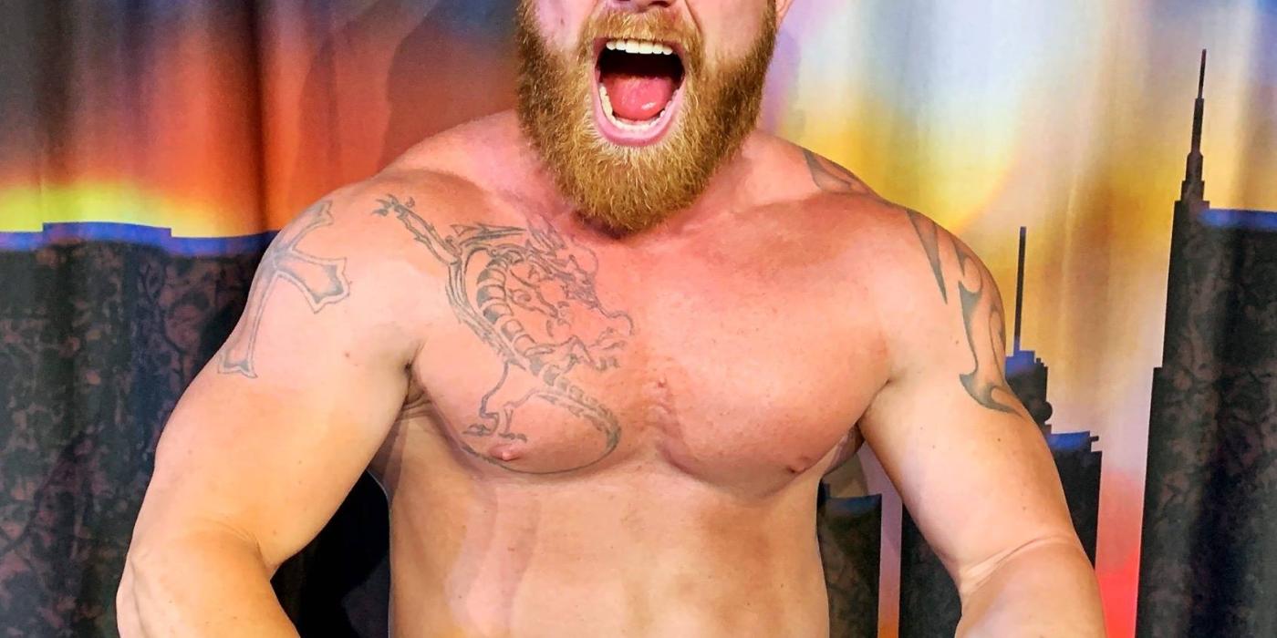 Ginger Billy wearing a baseball hat backwards, shirtless, displaying tattoos, doing a  muscle man pose holding a microphone
