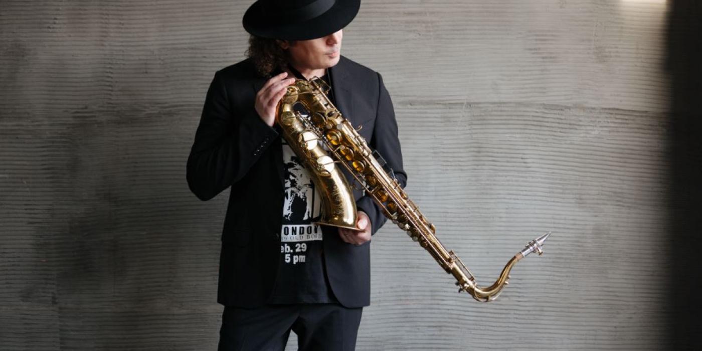 A man with shoulder length curly hair stands against a gray concrete wall, dressed in black with a black fedora. He holds his saxphone in his hands at an angle 