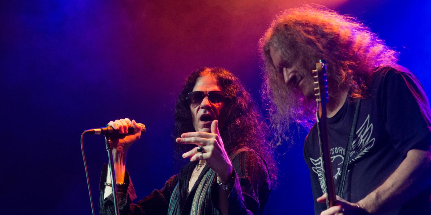 A man with long hair and sunglasses, sings into a microphone while a man with long hair plays a guitar next to him