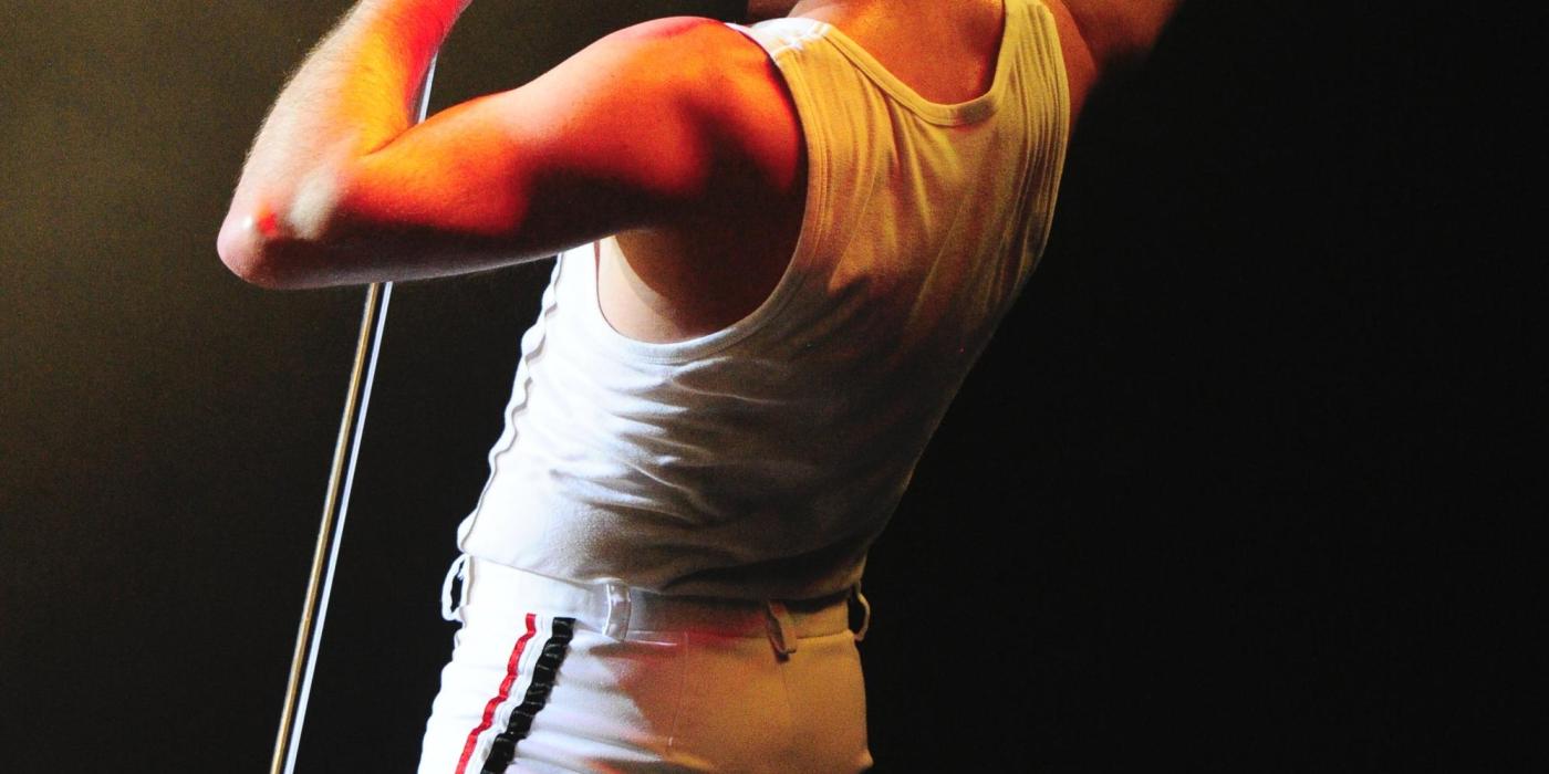 A man dressed in a white sleeveless shirt sings into a microphone with one arm raised