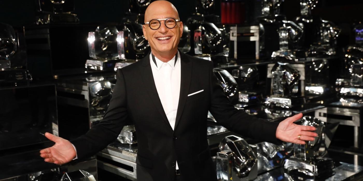 A white man with no hair standing on stage wearing a black suit and white shirt and glasses