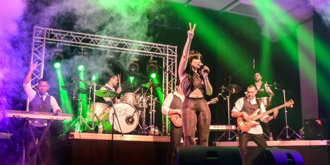 A full band plays on a stage with green and purple lights overhead. A woman stands at the front with one arm raised and a microphone in the other, singing.