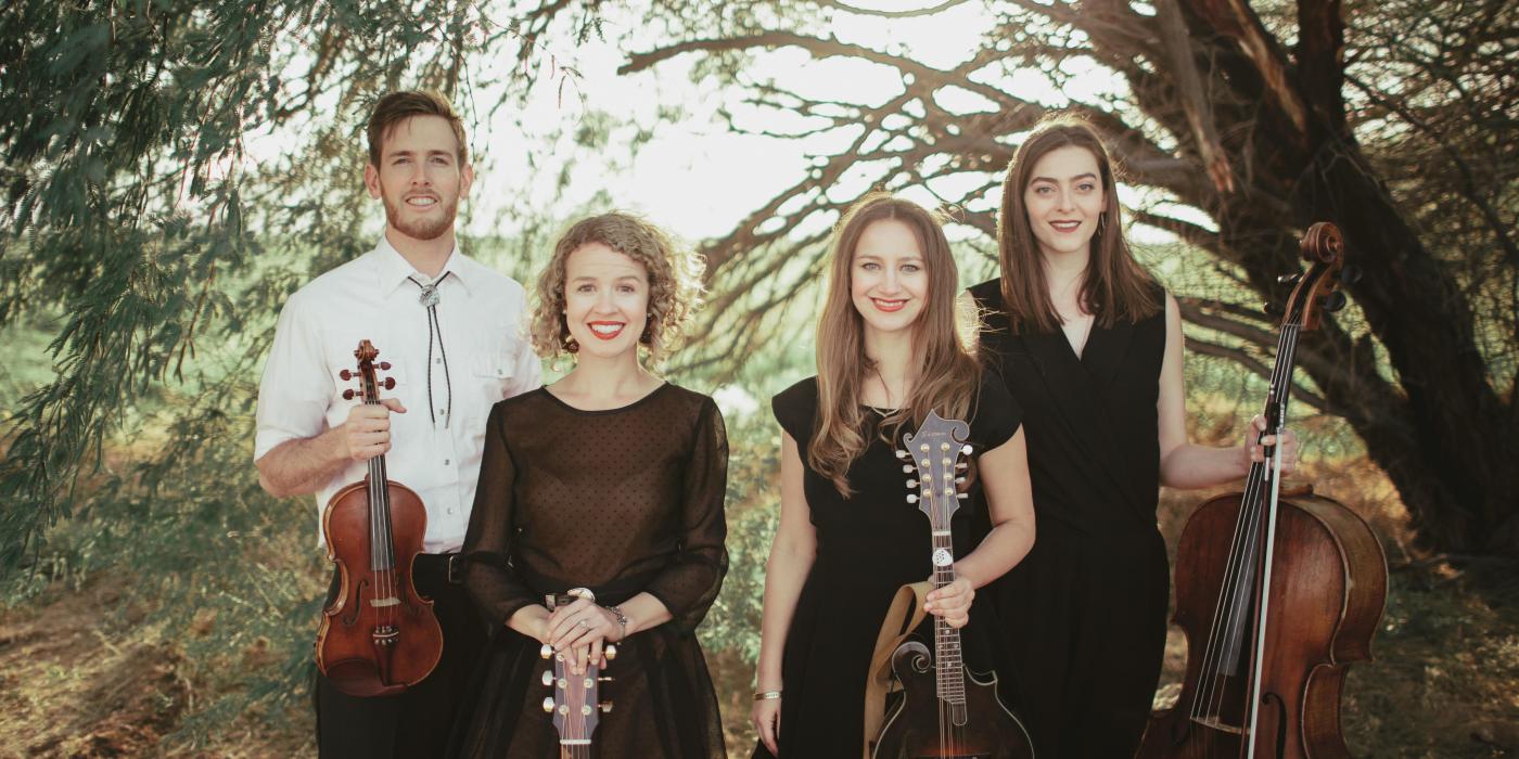 A man and three women look at the camera, string instruments in their hands with trees creating a green background