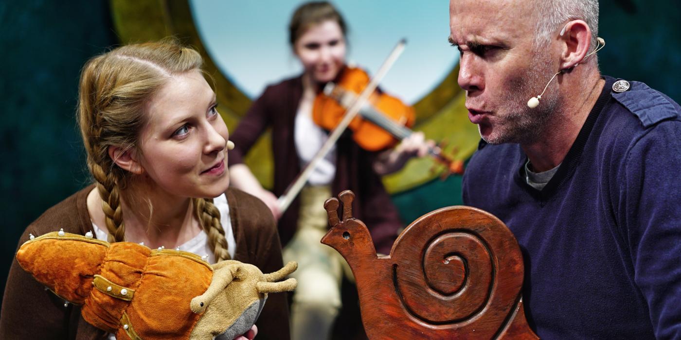 A young woman and an older man both hold small snail sculptures in their hand and talk while another woman plays violin behind them