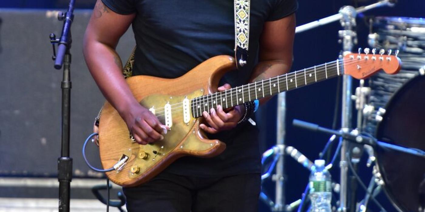 A man plays guitar on stage, his guitar is golden, he is wearing all black and he has tight curly hair colored platinum blond