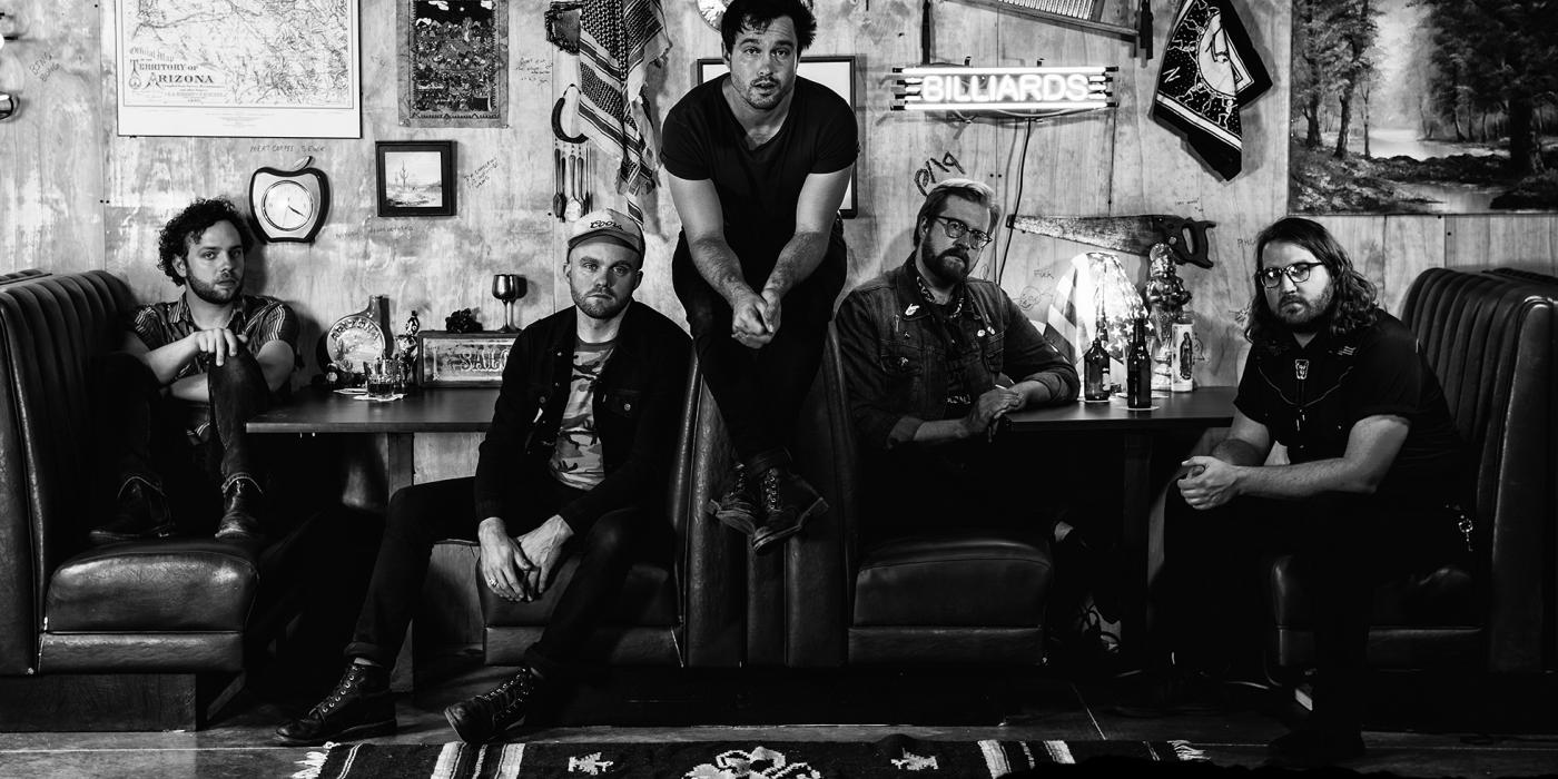In a black-and-white photo, five male band members of Jared and The Mill sit in a booth at a diner.