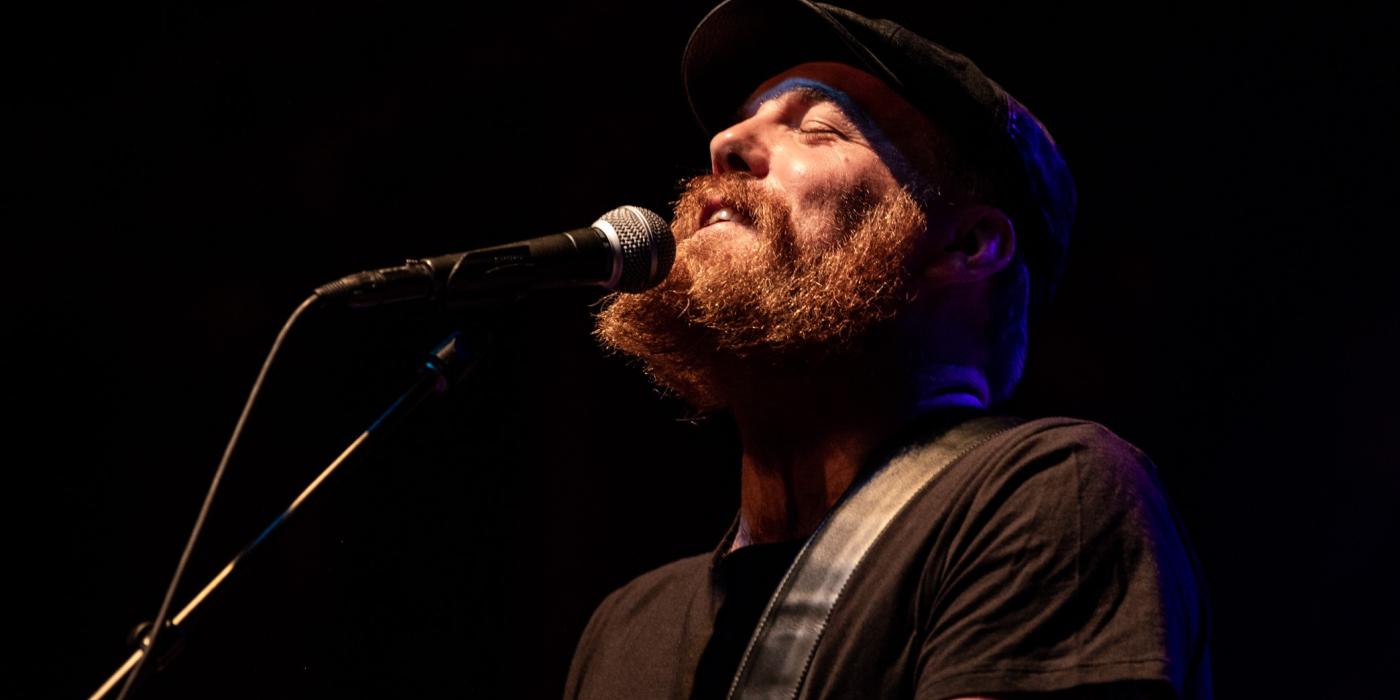 A bearded man dressed in black sings into a microphone and plays guitar