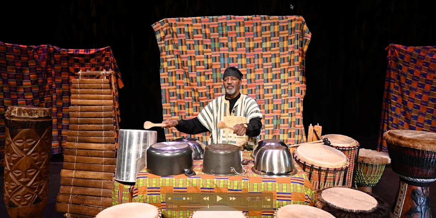 Percussionist Keith Johnson sits in traditional African clothing, playing a variety of percussion instruments including African drums, pots and pans.