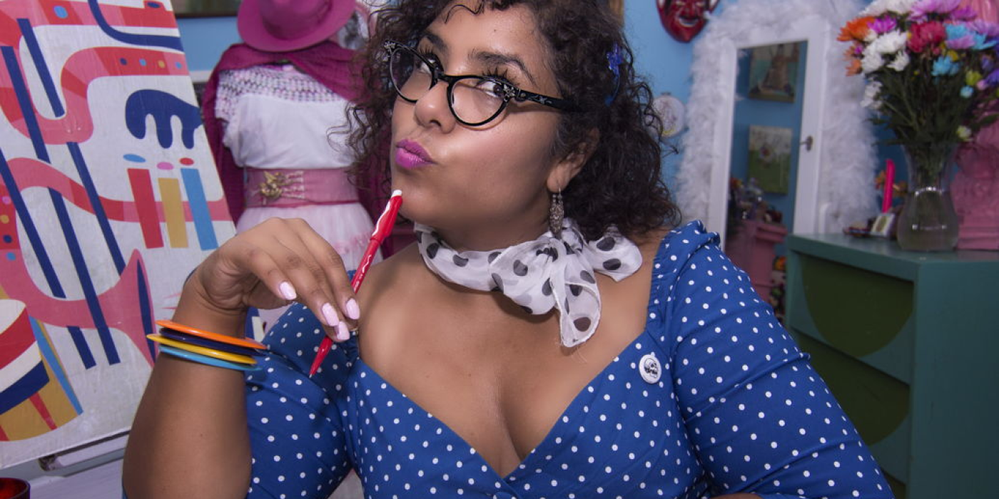 La Marisoul sitting at a desk in a colorful blue dress writing a letter and looking off to the side with a silly look on her face