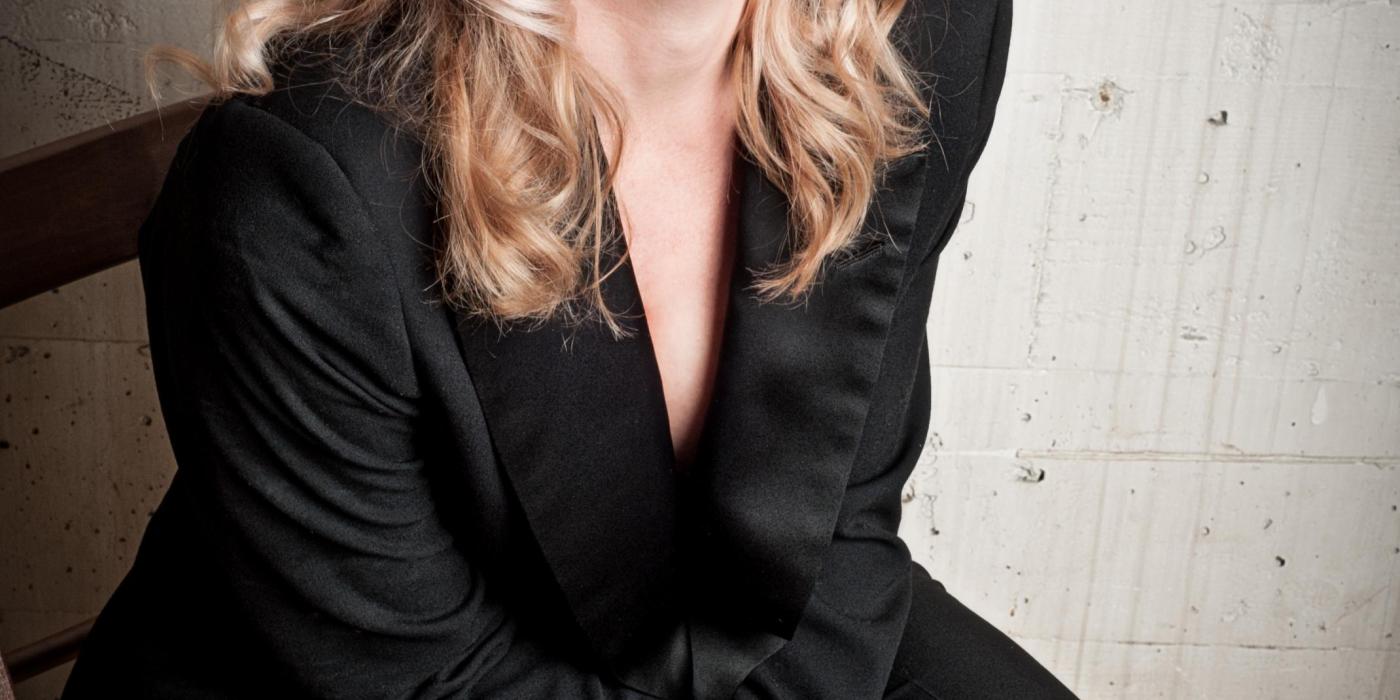 A blond woman sits in a black tailored suit, looking at the camera with sultry eyes